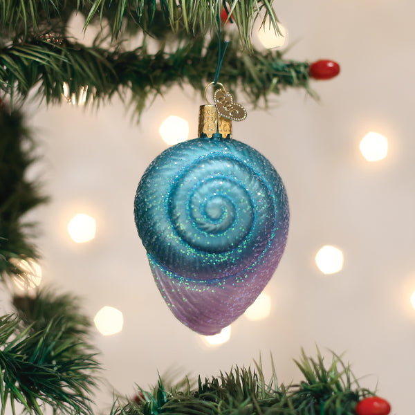 A blue and purple spiral seashell ornament hangs from a Christmas tree branch, surrounded by green pine needles and red berries, with soft white lights glowing in the background.