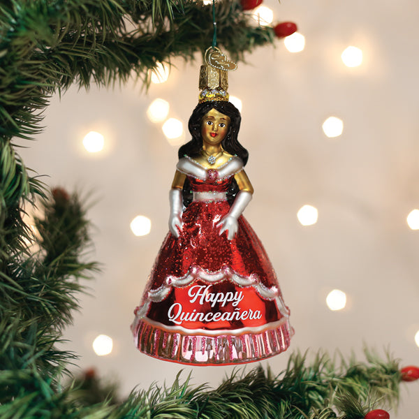 A Christmas tree ornament shaped like a young woman in a red glittery dress with the words Happy QuinceaÃ±era hangs from a green tree branch, with blurred lights in the background.
