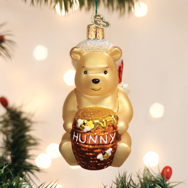 A Christmas tree ornament shaped like a golden bear holding a honey pot labeled HUNNY, surrounded by festive lights and green pine branches.